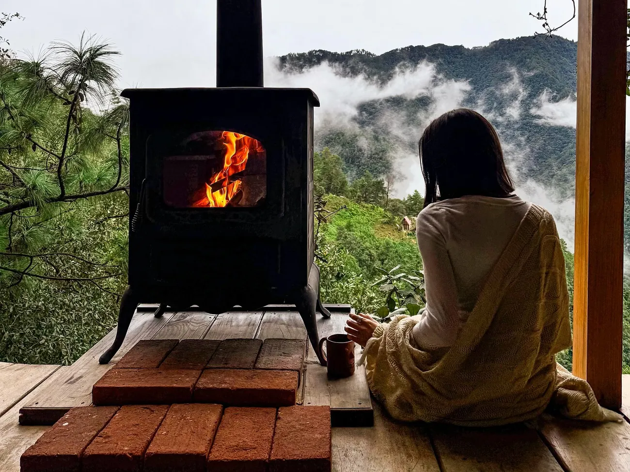 Una persona envuelta en una cobija junto a la estufa de leña, con nubes en el valle.